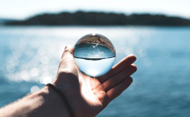 An outstretched hand over a waterbody holds an orb reflecting clear water. 